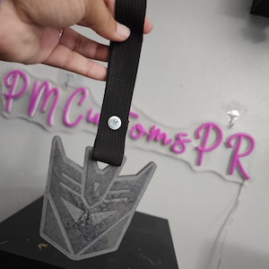 May include: A gray, textured pendant in the shape of a Transformers Decepticon symbol, attached to a black strap. The pendant is suspended against a light gray background. The strap is secured with a silver rivet.