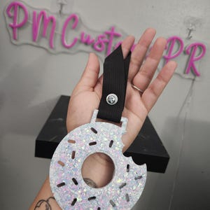 May include: A glittery, donut-shaped keychain with a bite taken out of it. The keychain is white with iridescent glitter and a black strap. The text "PM Customs PR" is visible in the background.