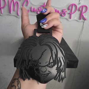 May include: A black acrylic car air freshener charm featuring a detailed engraved illustration of the Chucky character with a stitched face and messy hair, attached to a black elastic hanging loop for display in a vehicle or interior space.