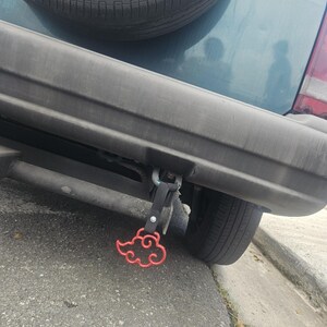 May include: A red cloud-shaped charm attached to a black fabric strap hanging from the rear bumper of a dark teal vehicle. The charm features a stylized, swirling cloud design with a metallic finish, suspended by a metal bolt and a black nylon loop.