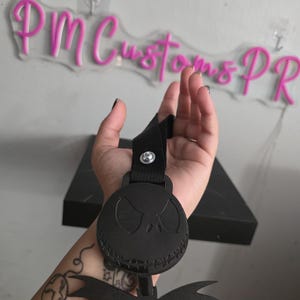 May include: Black wristband featuring a circular Jack Skellington design. The wristband has a black strap and a decorative bow. The background includes a neon sign that reads "PM Customs PR".