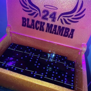May include: A gold-colored box with the words "24 BLACK MAMBA" and a wing design. Inside the box are black dominoes with blue dots. The box is open, revealing the dominoes.