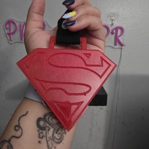 May include: A red Superman logo pendant with a black strap. The pendant is diamond-shaped with the iconic "S" symbol. The pendant is held up against a gray wall.
