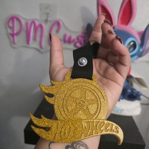 May include: A gold glittered Hot Wheels logo with a black strap and a silver screw. The logo features a wheel design with flames and the words "Hot Wheels".