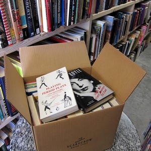 May include: A cardboard box filled with books, including titles like "Playing Period Plays" and "BOYLE." The box is labeled "SCARECROW BOOK SHOP." Bookshelves filled with various titles are visible in the background, creating a bookstore setting.