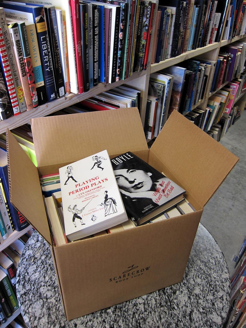 May include: A cardboard box filled with books, including titles like "Playing Period Plays" and "BOYLE." The box is labeled "SCARECROW BOOK SHOP." Bookshelves filled with various titles are visible in the background, creating a bookstore setting.