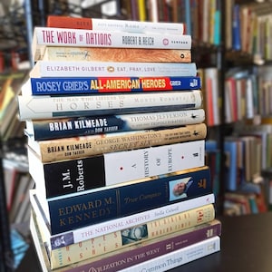 May include: A stack of hardcover books with various titles, including "The Work of Nations" and "Eat, Pray, Love." The spines display a range of colors, with the books arranged on a dark surface, suggesting a library or bookstore setting.
