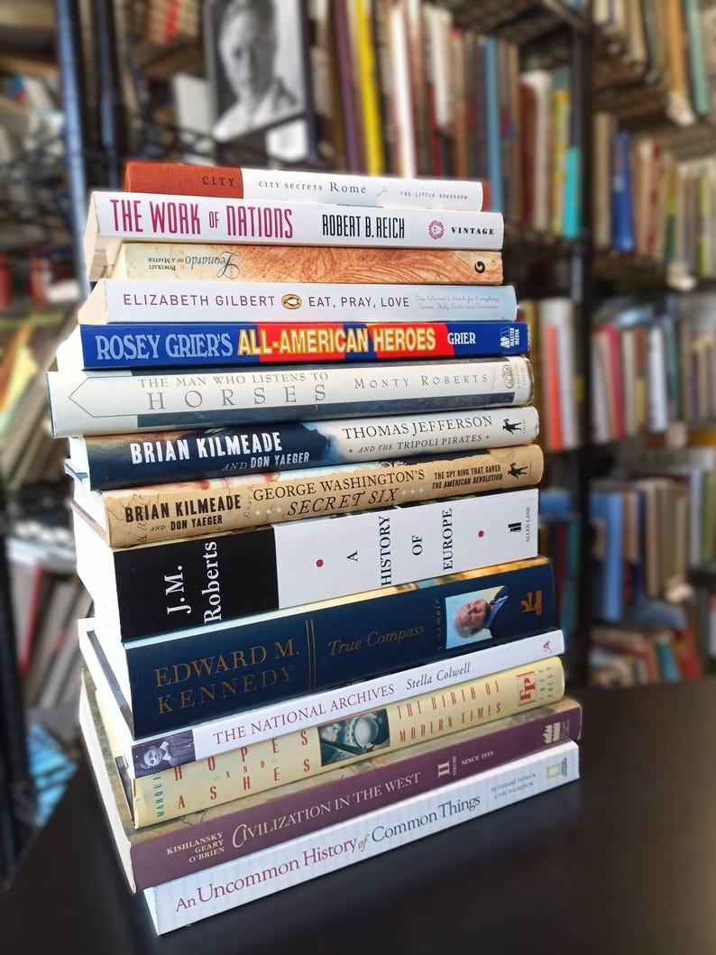 May include: A stack of hardcover books with various titles, including "The Work of Nations" and "Eat, Pray, Love." The spines display a range of colors, with the books arranged on a dark surface, suggesting a library or bookstore setting.