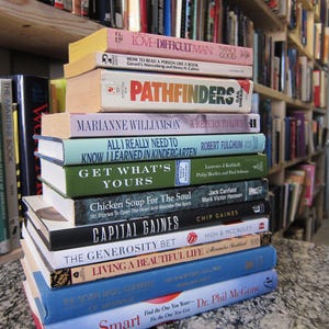 May include: A stack of hardcover books with various titles and colors. Titles include "Love Smart", "Capital Gaines", and "Chicken Soup for the Soul". The books are stacked on a speckled surface, with a bookshelf in the background.