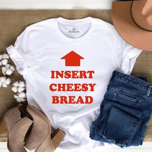 May include: White t-shirt with the text "INSERT CHEESY BREAD" in red, with a red arrow pointing upwards. The shirt is paired with blue jeans, tan boots, and a brown hat. The shirt is on a burlap surface.