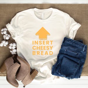 May include: White t-shirt with the text "INSERT CHEESY BREAD" in orange, with an orange arrow graphic. The shirt is paired with blue jeans and tan ankle boots with block heels. Cotton sprigs are arranged on the side.