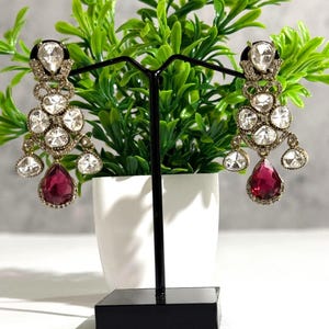 May include: A pair of ornate chandelier earrings. Each earring features multiple clear, sparkling stones and a large, teardrop-shaped, ruby-red stone at the bottom. The earrings are displayed on a black stand in front of a green plant.