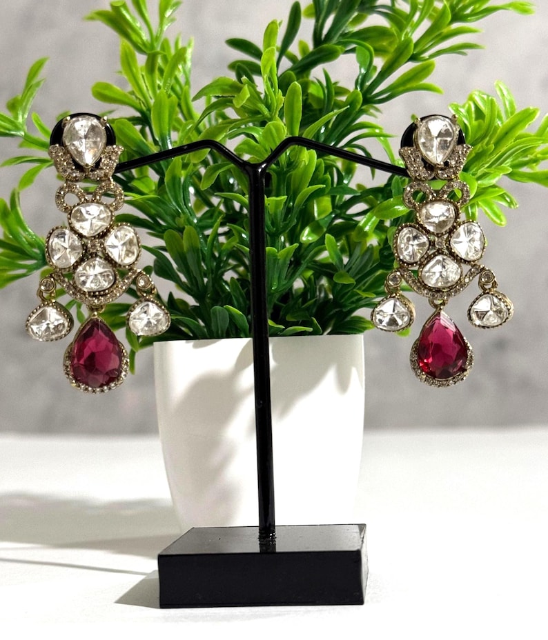 May include: A pair of ornate chandelier earrings. Each earring features multiple clear, sparkling stones and a large, teardrop-shaped, ruby-red stone at the bottom. The earrings are displayed on a black stand in front of a green plant.