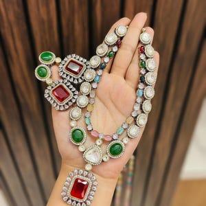May include: A multi-strand necklace and matching earrings set. The jewelry features large, colorful gemstones in red, green, and clear, surrounded by smaller, sparkling stones. The necklace has a large, rectangular pendant.