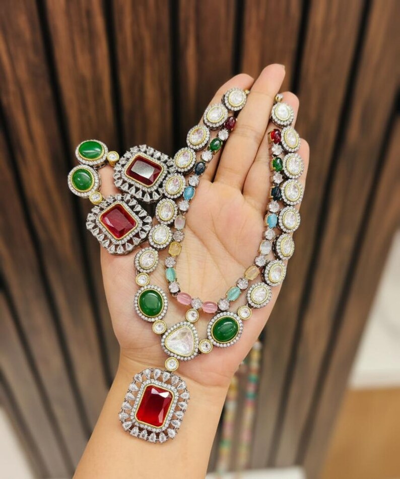 May include: A multi-strand necklace and matching earrings set. The jewelry features large, colorful gemstones in red, green, and clear, surrounded by smaller, sparkling stones. The necklace has a large, rectangular pendant.