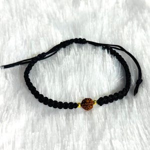 Peut inclure: Bracelet noir réglable avec une perle centrale marron et des détails dorés. Le bracelet est fait de cordon noir tressé et est réglable pour un ajustement confortable. Le bracelet est présenté sur un fond blanc texturé.
