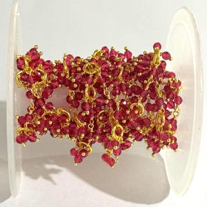 Puede incluir: Un carrete de cadena de piedras preciosas rojas con eslabones de metal dorados. La cadena está hecha de pequeñas cuentas redondas de color rojo intenso. Los eslabones dorados conectan las cuentas, creando un diseño delicado e intrincado. La cadena está enrollada alrededor de un carrete de plástico transparente.