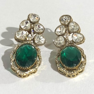 May include: A pair of elaborate earrings. Each earring features a cluster of clear, faceted stones above a large, oval emerald-green stone. The earrings have a gold-coloured metal setting with intricate detailing, creating a luxurious appearance.
