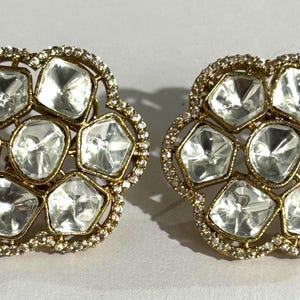 Könnte beinhalten: Ein Paar goldfarbene Ohrstecker. Jeder Ohrring hat ein Blumenmuster mit klaren, facettierten Edelsteinen in einer Anordnung, umgeben von kleineren, klaren Steinen. Die Ohrringe sind vor einem weißen Hintergrund platziert.