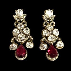 May include: A pair of ornate gold-toned earrings with a cascading design. Each earring features multiple clear, sparkling stones and a large, teardrop-shaped, deep red gemstone at the bottom. The earrings are set against a black background.