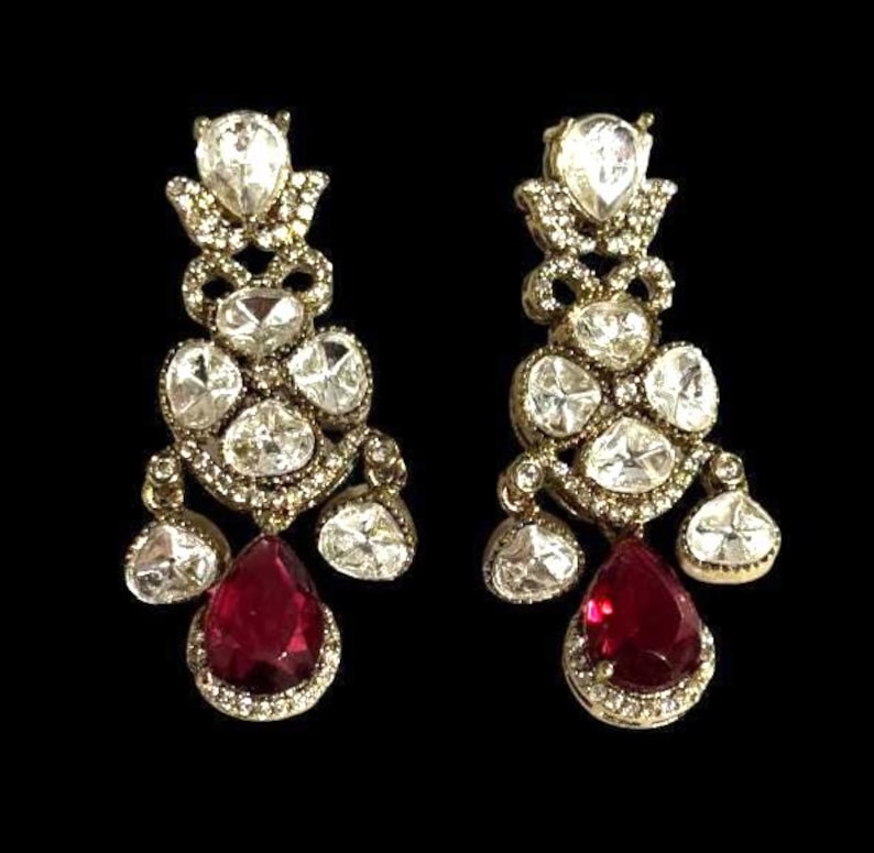 May include: A pair of ornate gold-toned earrings with a cascading design. Each earring features multiple clear, sparkling stones and a large, teardrop-shaped, deep red gemstone at the bottom. The earrings are set against a black background.