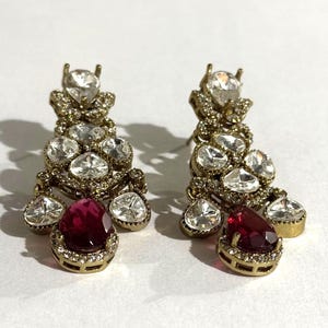 May include: A pair of gold-toned chandelier earrings. Each earring features a cluster of clear, sparkling stones and a large, teardrop-shaped, ruby-red stone at the bottom. The earrings have a vintage or antique aesthetic.