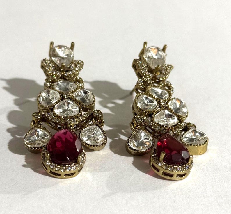 May include: A pair of gold-toned chandelier earrings. Each earring features a cluster of clear, sparkling stones and a large, teardrop-shaped, ruby-red stone at the bottom. The earrings have a vintage or antique aesthetic.
