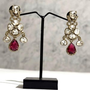 May include: A pair of chandelier earrings with a gold-tone setting. Each earring features multiple clear, round-cut stones and a large, teardrop-shaped, ruby-red stone at the bottom. The earrings are displayed on a black stand.