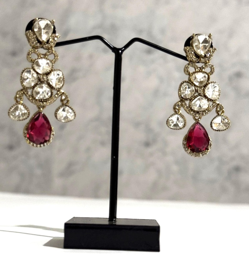 May include: A pair of chandelier earrings with a gold-tone setting. Each earring features multiple clear, round-cut stones and a large, teardrop-shaped, ruby-red stone at the bottom. The earrings are displayed on a black stand.