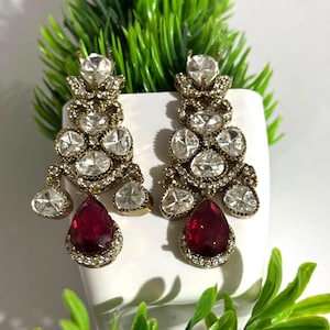 May include: A pair of ornate chandelier earrings. The earrings feature multiple clear, sparkling stones and a large, teardrop-shaped, ruby-red stone at the bottom. The metal has a gold tone. The earrings are displayed against a white background with green foliage.