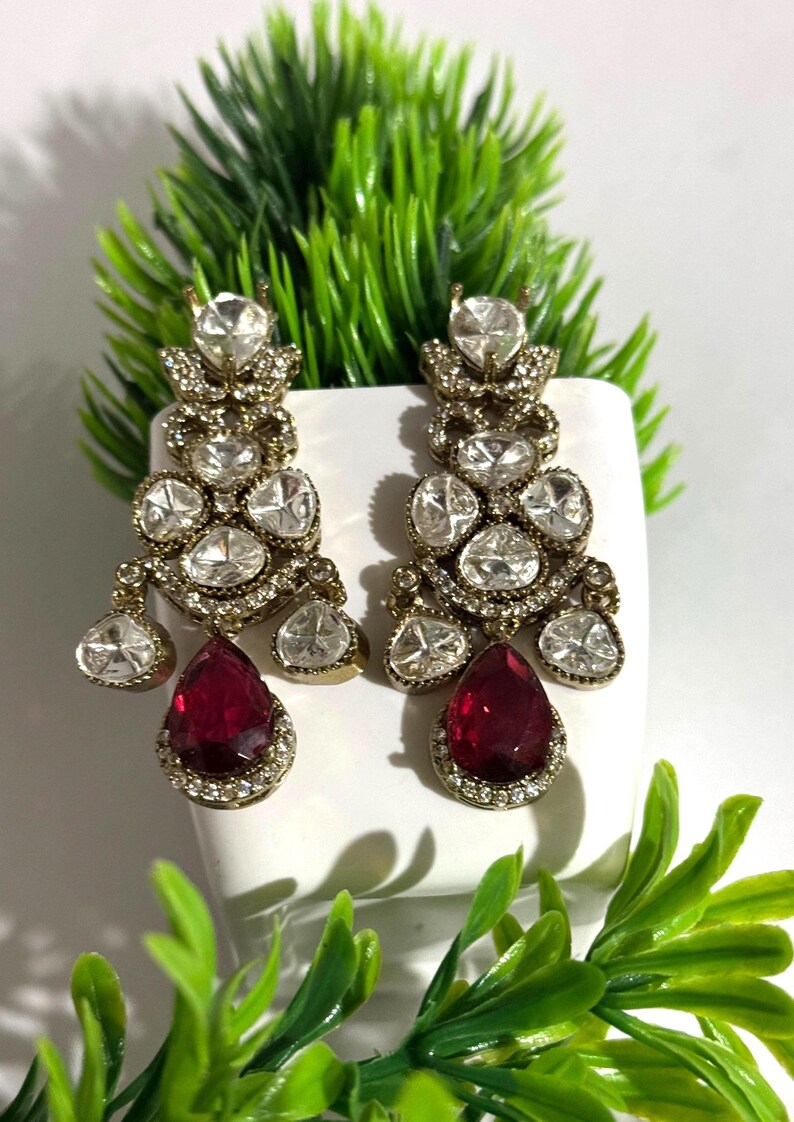 May include: A pair of ornate chandelier earrings. The earrings feature multiple clear, sparkling stones and a large, teardrop-shaped, ruby-red stone at the bottom. The metal has a gold tone. The earrings are displayed against a white background with green foliage.