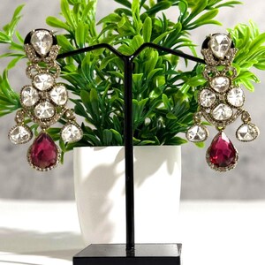 May include: A pair of ornate chandelier earrings. Each earring features multiple clear, round-cut stones and a large, teardrop-shaped, ruby-red stone at the bottom. The earrings are displayed on a black stand against a backdrop of green foliage.