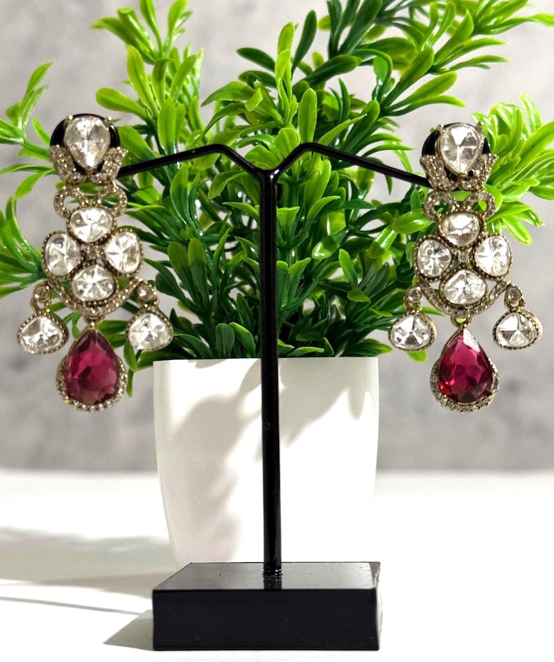 May include: A pair of ornate chandelier earrings. Each earring features multiple clear, round-cut stones and a large, teardrop-shaped, ruby-red stone at the bottom. The earrings are displayed on a black stand against a backdrop of green foliage.