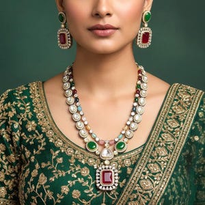 May include: A woman wearing a green and gold embroidered top and a multi-layered necklace with matching earrings. The necklace features a large, square, red gemstone pendant and is adorned with white, green, and other colorful gemstones. The earrings have a similar design.