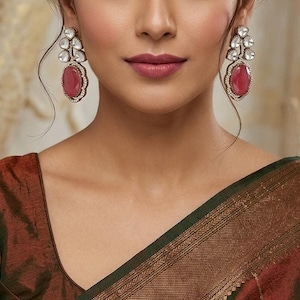 May include: A woman wearing large, ornate earrings with a ruby-red gemstone center and clear, sparkling accents. The earrings are set in a silver-toned metal. She is wearing a rust-colored blouse with a green and gold-toned trim.