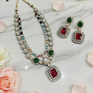 May include: A multi-strand necklace and matching earrings set. The necklace features a large, square, red gemstone pendant and is adorned with various colored gemstones and clear crystals. The earrings have a similar design with red and green gemstones.