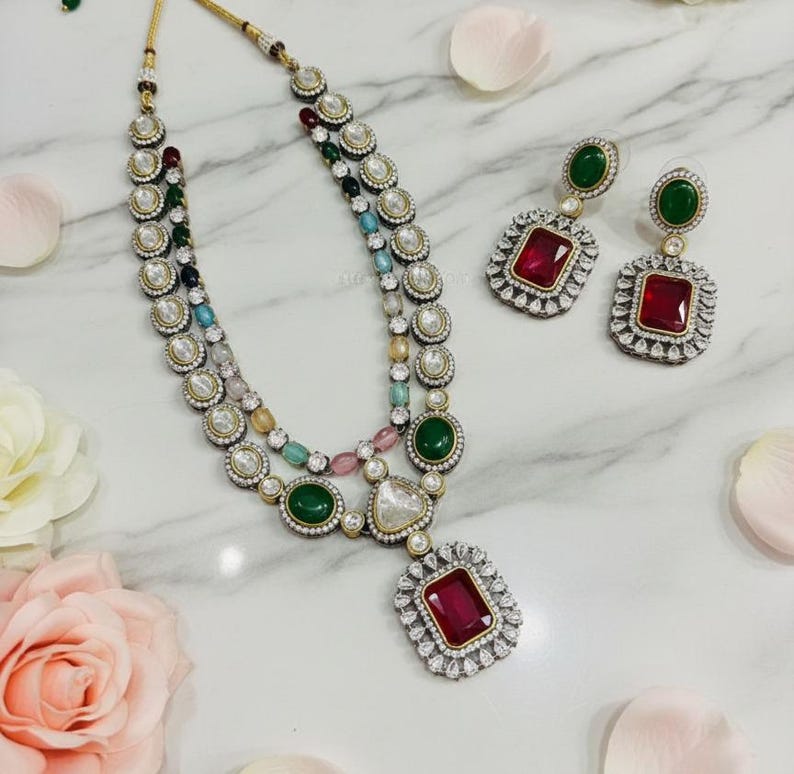 May include: A multi-strand necklace and matching earrings set. The necklace features a large, square, red gemstone pendant and is adorned with various colored gemstones and clear crystals. The earrings have a similar design with red and green gemstones.