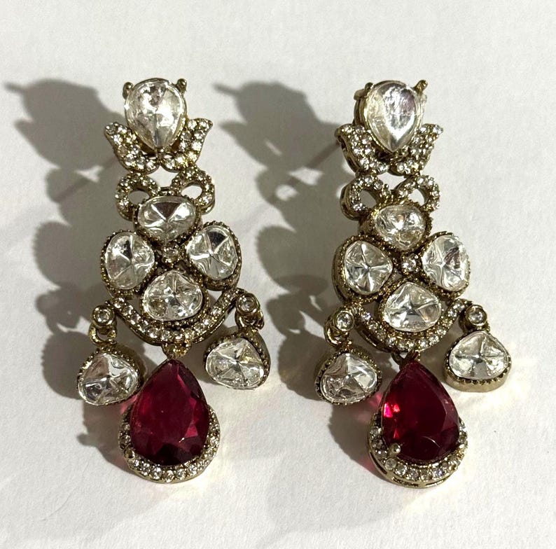 May include: A pair of ornate drop earrings. Each earring features a teardrop-shaped, deep red gemstone at the bottom, surrounded by smaller clear stones. The earrings have a gold-colored metal setting and are adorned with multiple clear, sparkling stones.