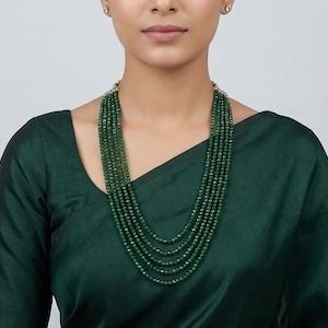 May include: A layered necklace featuring multiple strands of small, dark green beads. The necklace is worn by a person wearing a dark green top. The beads appear to be evenly sized and closely spaced.