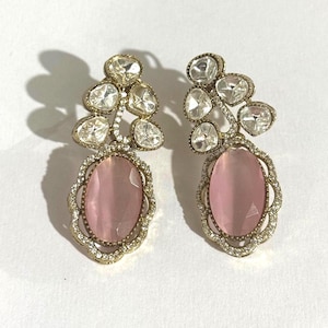 Può includere: Un paio di orecchini pendenti color oro. Ogni orecchino presenta un gruppo di pietre chiare e sfaccettate nella parte superiore, collegato a una pietra rosa ovale. La pietra rosa è circondata da una cornice decorativa con piccole pietre chiare.
