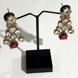 May include: A pair of gold-toned chandelier earrings with clear and red gemstones. The earrings are displayed on a black stand against a white background. The earrings have multiple clear, round-cut stones and a large, teardrop-shaped red stone.