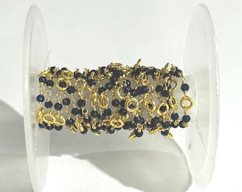 May include: A spool of gold-colored chain with small, dark blue beads. The chain is wound around a clear plastic spool, creating a layered effect. The beads are evenly spaced along the chain.