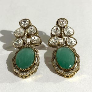Könnte beinhalten: Ein Paar verzierter Ohrringe mit klaren, facettierten Steinen und einem großen, ovalen, teal-farbenen Stein. Die Ohrringe haben eine goldfarbene Metalleinfassung mit aufwendigen Details und kleinen, klaren Akzentsteinen um den teal-farbenen Stein.