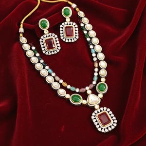 May include: A two-strand necklace and matching earrings set. The necklace features a large rectangular pendant with a red stone, surrounded by small white stones. The earrings have a similar design. The set includes green and white stones, and is set against a red background.