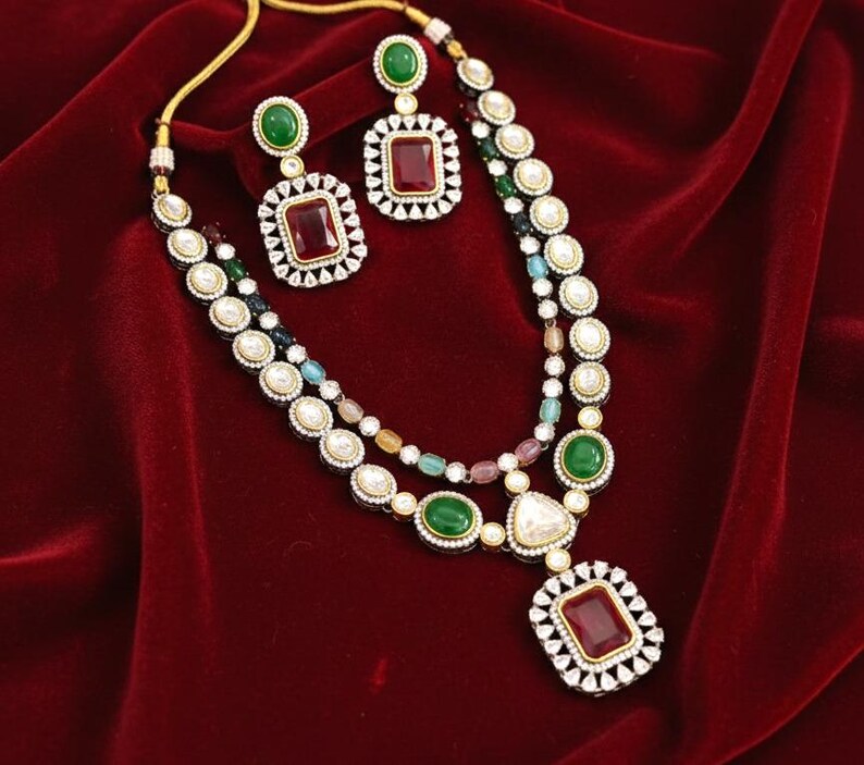 May include: A two-strand necklace and matching earrings set. The necklace features a large rectangular pendant with a red stone, surrounded by small white stones. The earrings have a similar design. The set includes green and white stones, and is set against a red background.