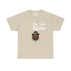Peut inclure: T-shirt beige avec l'inscription blanche "The Dogfather", représentant un bouledogue portant un chapeau et un collier clouté. Le t-shirt a un col rond et des manches courtes.