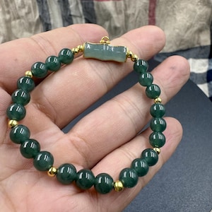 Puede incluir: Una pulsera de cuentas verde oscuro con detalles dorados. La pulsera presenta cuentas redondas y pulidas y pequeños espaciadores dorados. Una pieza rectangular de jade verde claro está centrada en la pulsera.
