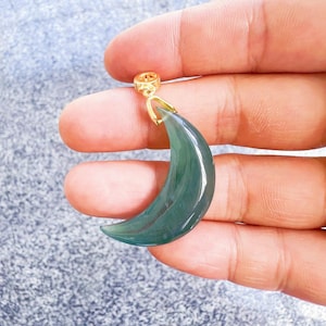 Puede incluir: Un colgante en forma de media luna de color verde jade translúcido, con una fianza dorada. La fianza está unida a la parte superior del colgante. El colgante está sujeto en una mano.
