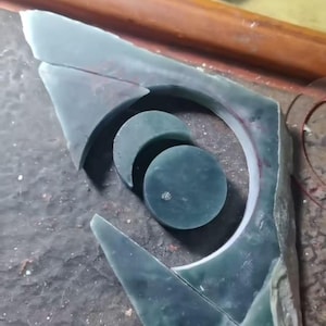May include: A collection of jade stone pieces in various shapes, including a crescent, a circle, and a large, irregular shape with a circular cutout. The jade is a deep teal color, and the pieces appear to be in the process of being crafted.