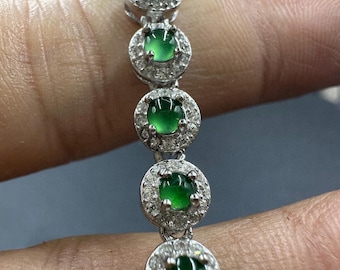 Op de afbeelding: Een zilveren armband met een reeks ronde, levendige groene edelstenen, elk omcirkeld door een halo van kleine, sprankelende, heldere stenen. De armband wordt gepresenteerd tegen een neutrale achtergrond, waardoor het ingewikkelde ontwerp van de sieraden wordt benadrukt.